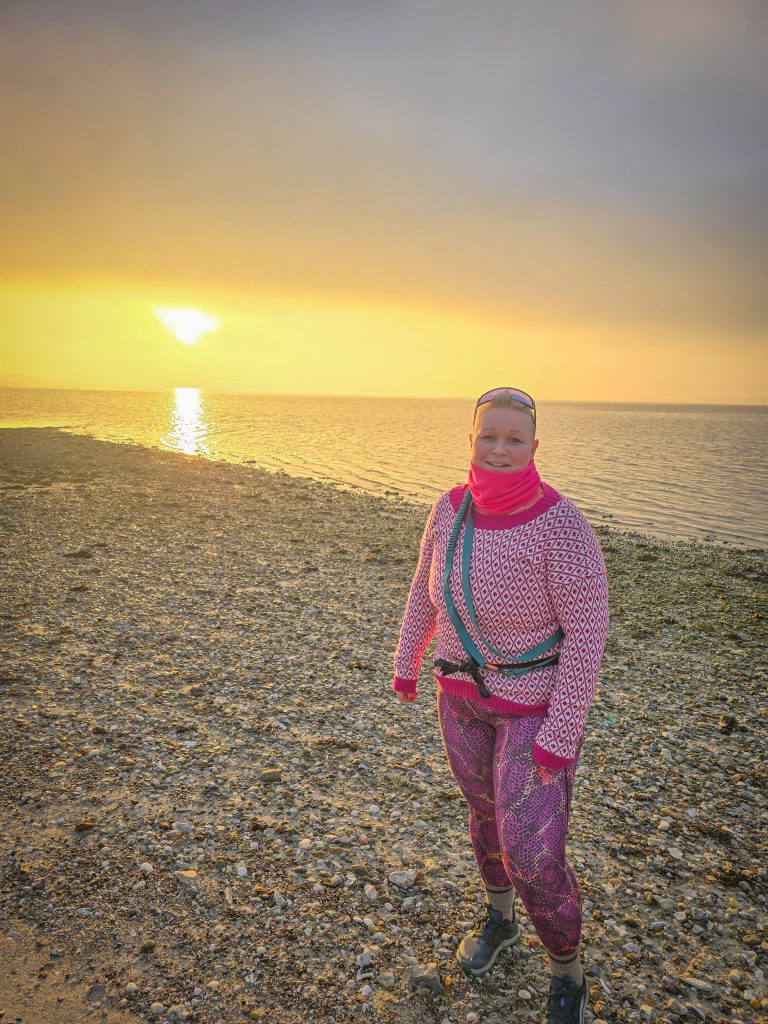 Woman wearing colourful outdoor clothing on a pebble beach during a golden sunset by the sea.