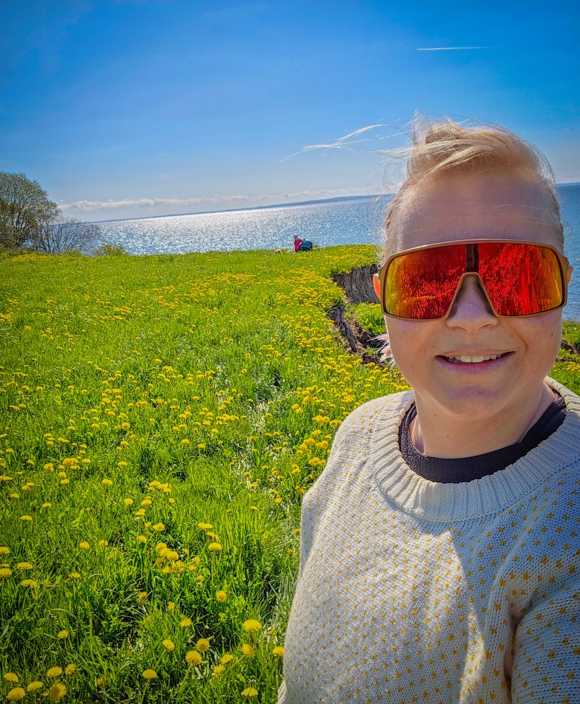 Woman hiking through a sunny coastal meadow filled with yellow wildflowers and the sea in the background.