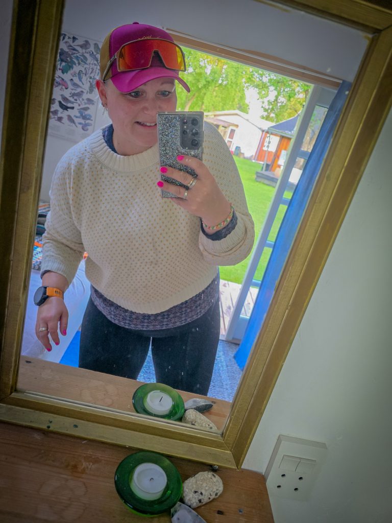 Woman wearing colourful outdoor clothing and sunglasses, taking a mirror selfie before heading outside.
