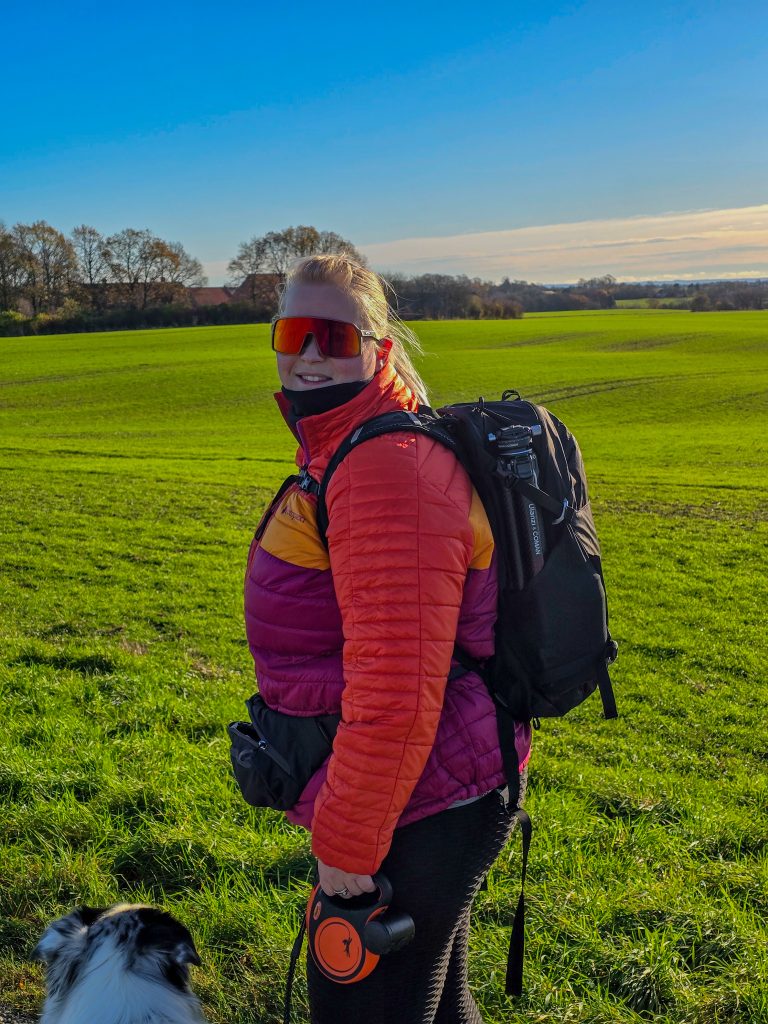 Woman wearing colourful outdoor clothing and a hiking backpack while walking with her dog in the countryside.