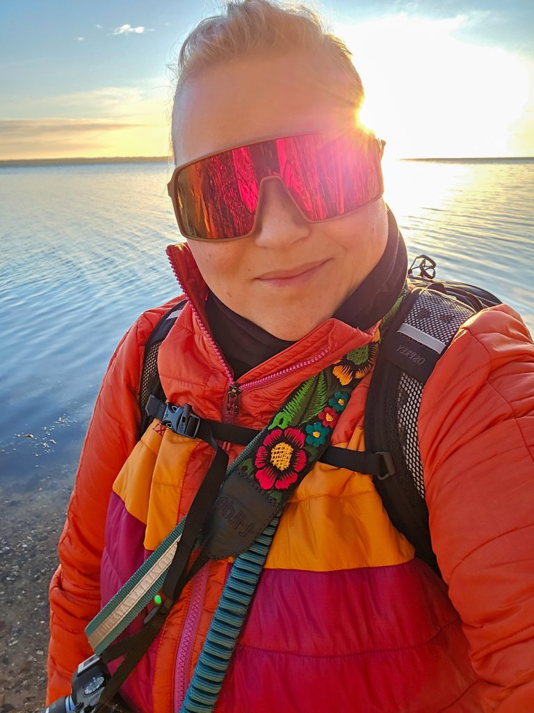 Woman wearing colourful outdoor clothing and a hiking backpack by the water at sunrise.