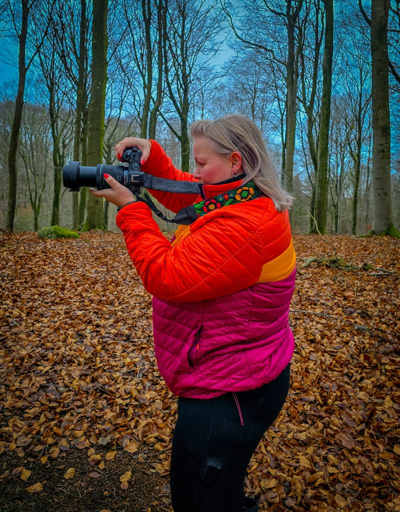Woman photographing in a forest while wearing a colourful outdoor jacket.