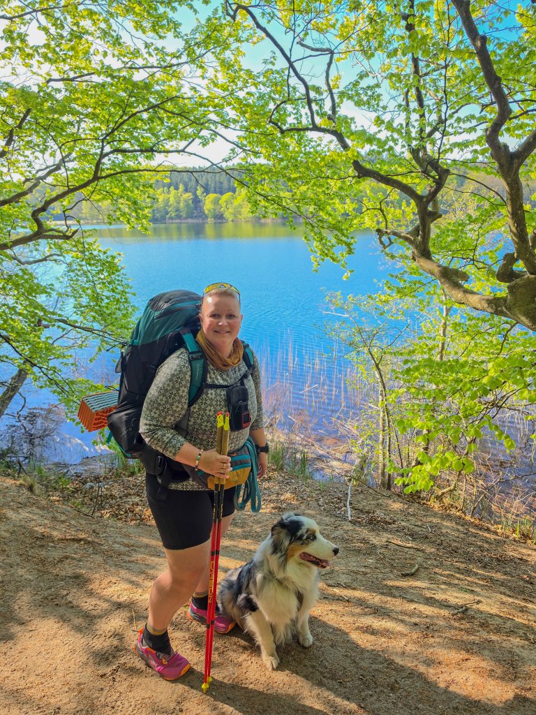 Woman backpacking with her Australian Shepherd beside a forest lake in Denmark.