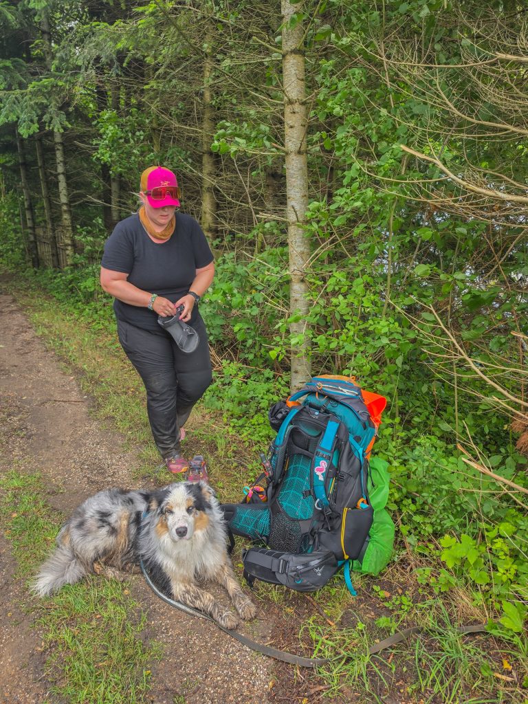 Woman taking a break with her muddy Australian Shepherd and backpack on a hiking trail in Denmark.