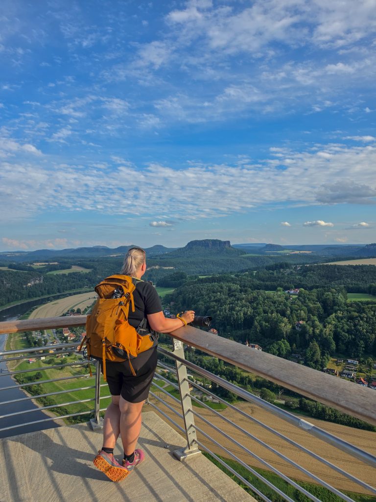 Woman with a hiking backpack and camera overlooking a mountain landscape from a viewpoint.