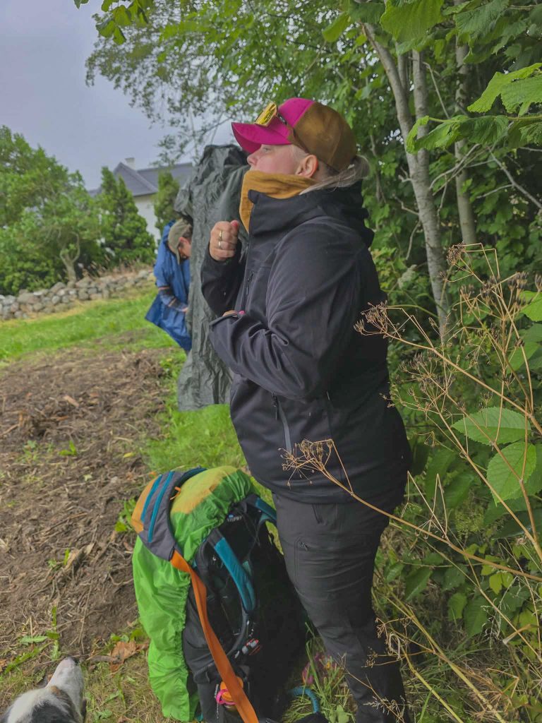 Woman in waterproof hiking clothes taking a break beside her backpack during a rainy hike in Denmark.