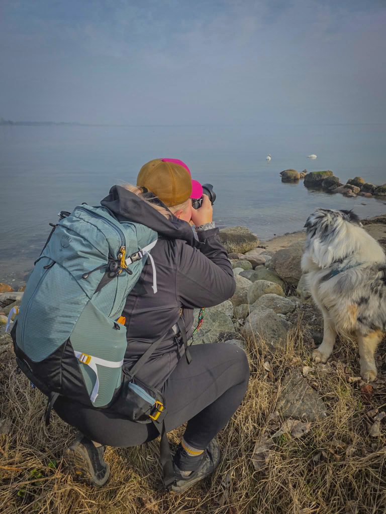 Woman photographing wildlife by the coast with her dog and hiking backpack.