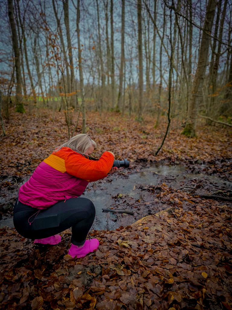 Woman in colourful outdoor clothing crouching by a forest stream while taking photographs.